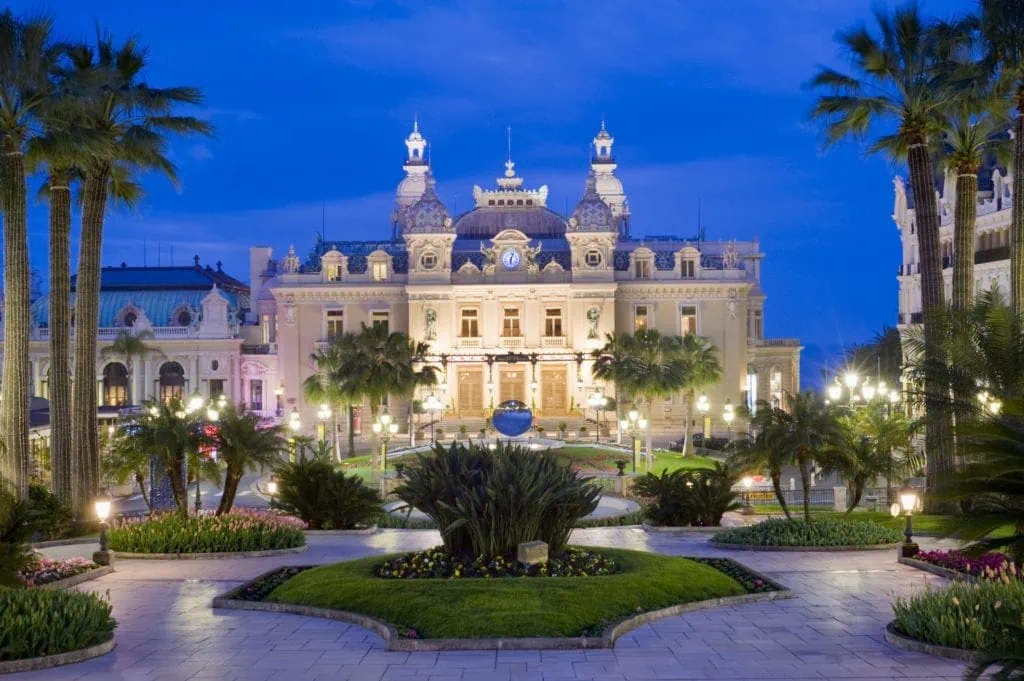 Elegant historic hotel with ornate architecture in night lighting, lush gardens and fountains in Monte Carlo.