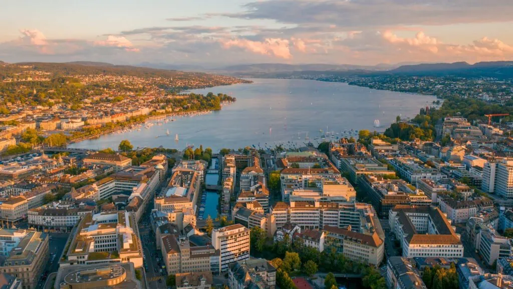 Panoramic view of Zurich with Lake Zurich, city skyline, and surrounding hills at sunset.