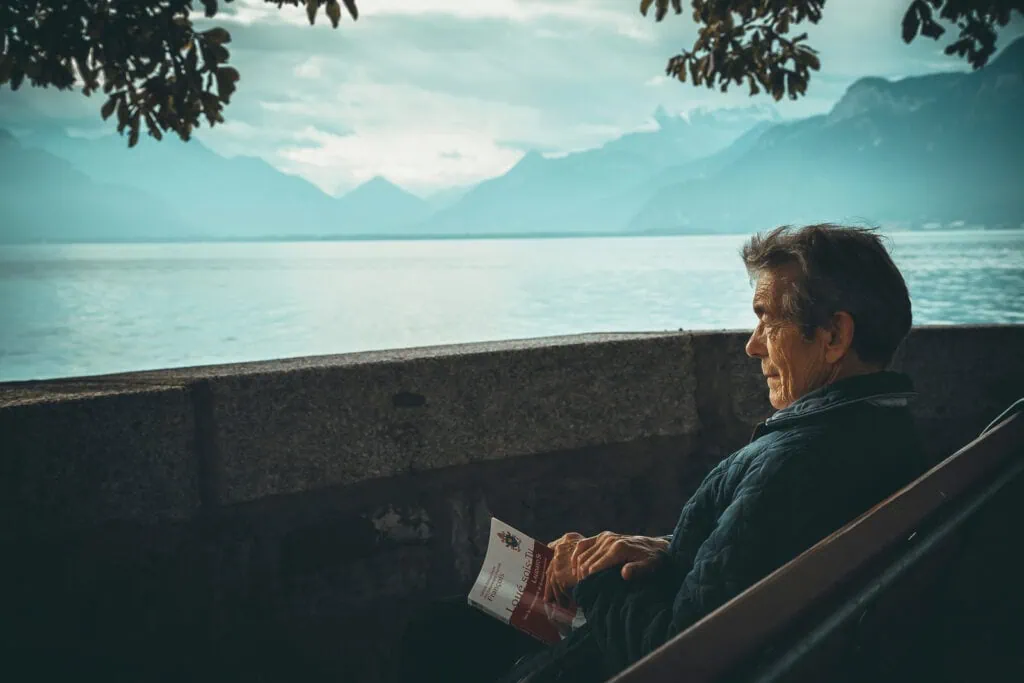 Serene lakeside view with elderly man reading, mountain backdrop, peaceful atmosphere.