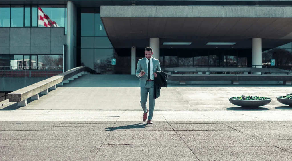 Young businessman walking towards office building, checking phone, in formal suit.