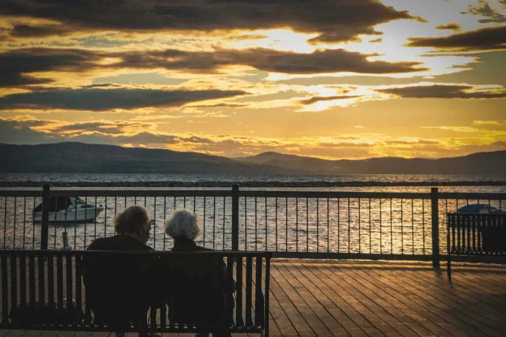 Serene sunset view of elderly couple on lakeside pier with boat and mountains, peaceful scenery.