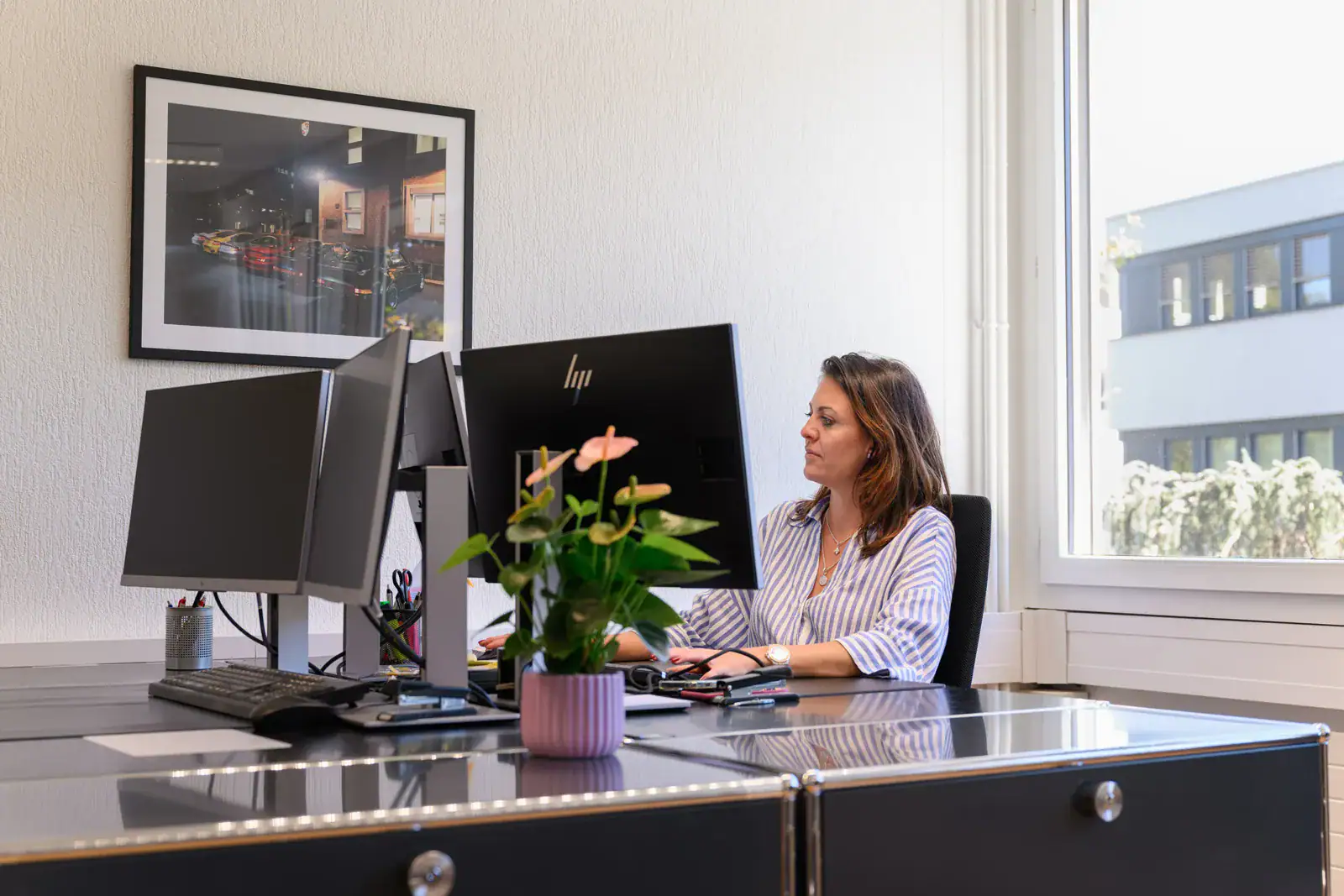 Modern woman working at an office desk with dual monitors and a potted plant.