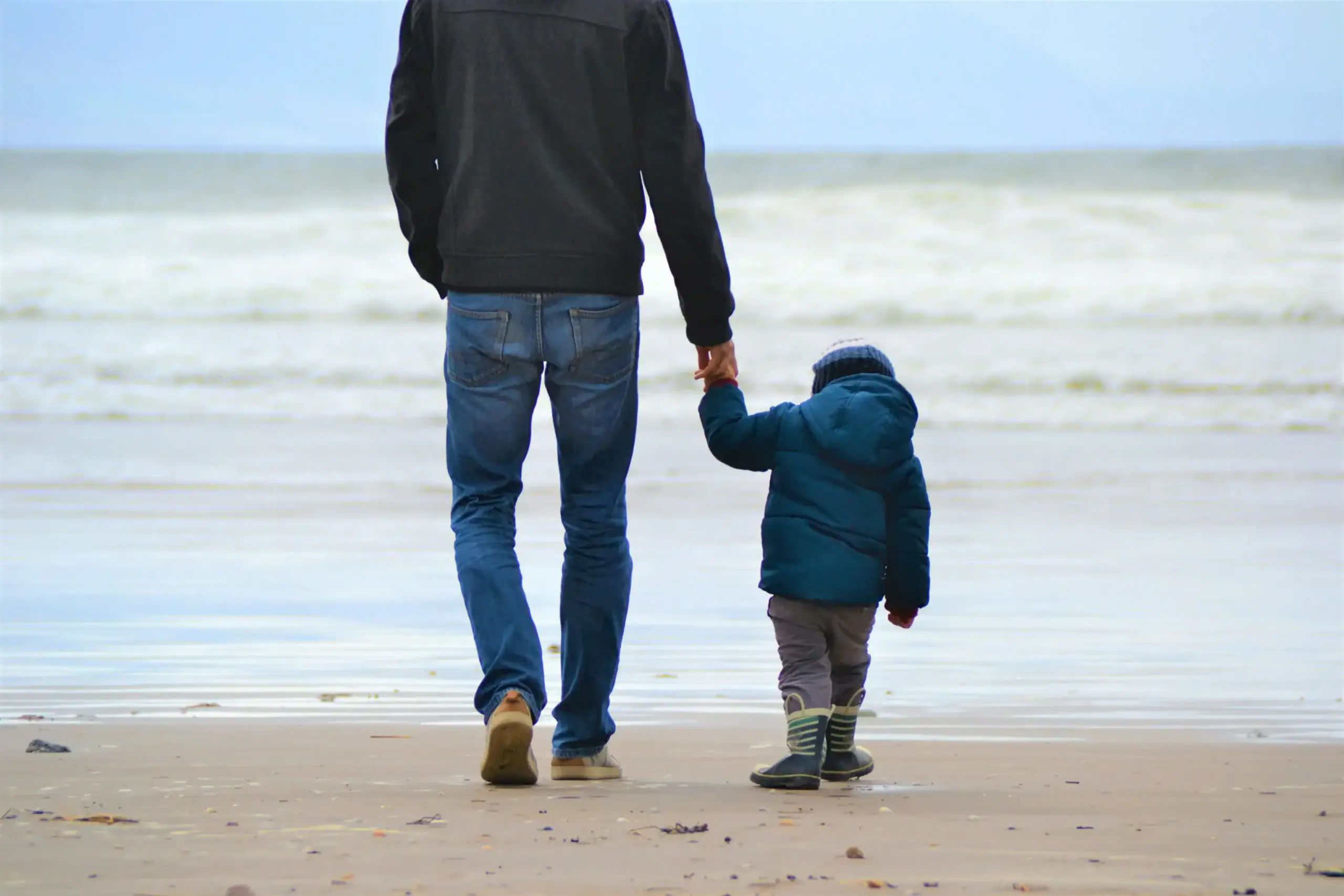 Father and child walking hand in hand on the beach with ocean in background.