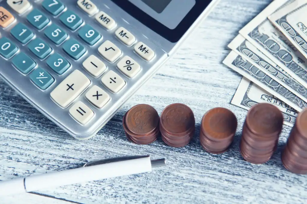 Calculator, stacked coins, pen, and US dollar bills on wooden surface for financial analysis.