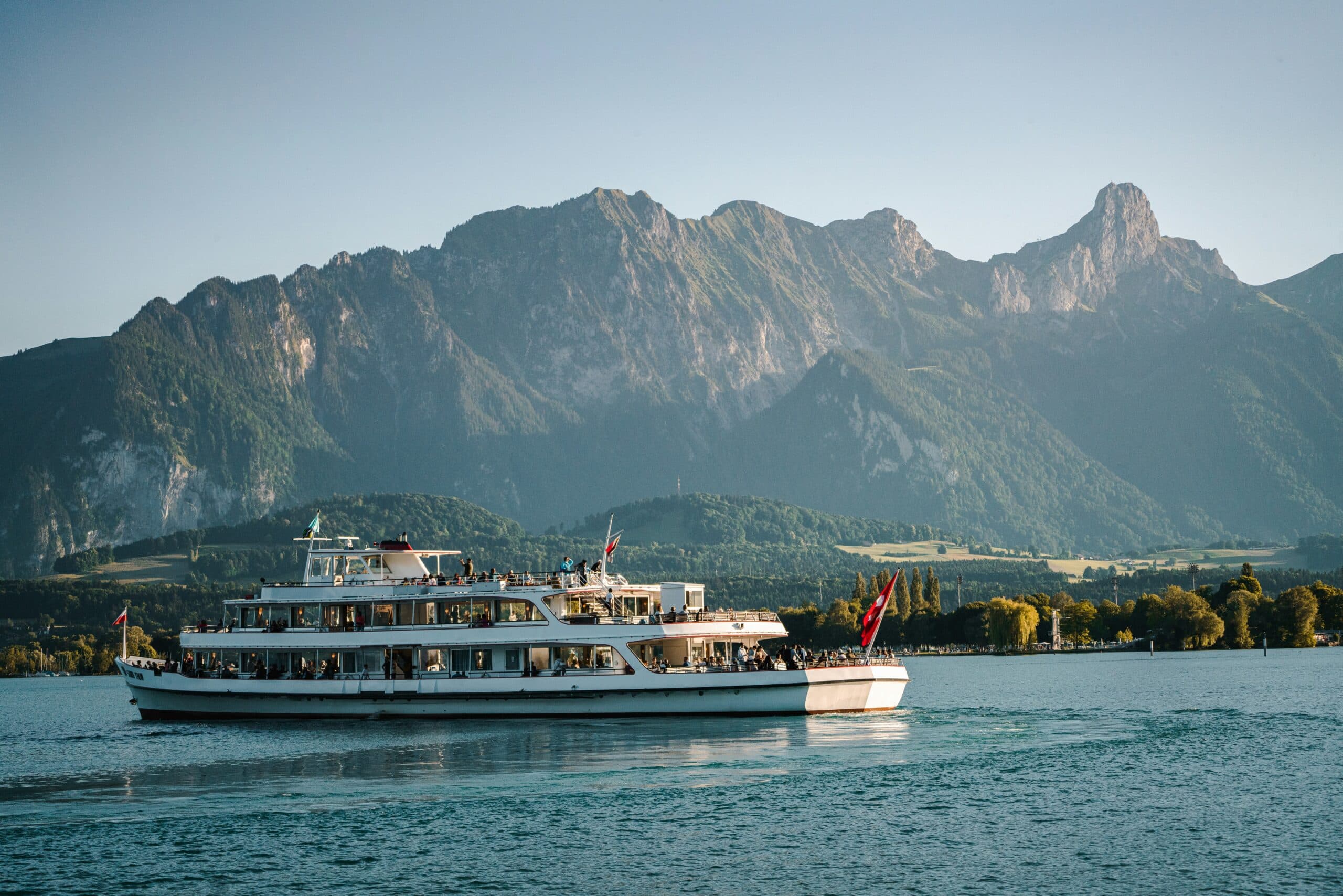White passenger boat flying Swiss flags on a calm lake, with wooded shores and a rugged mountain range in warm evening light