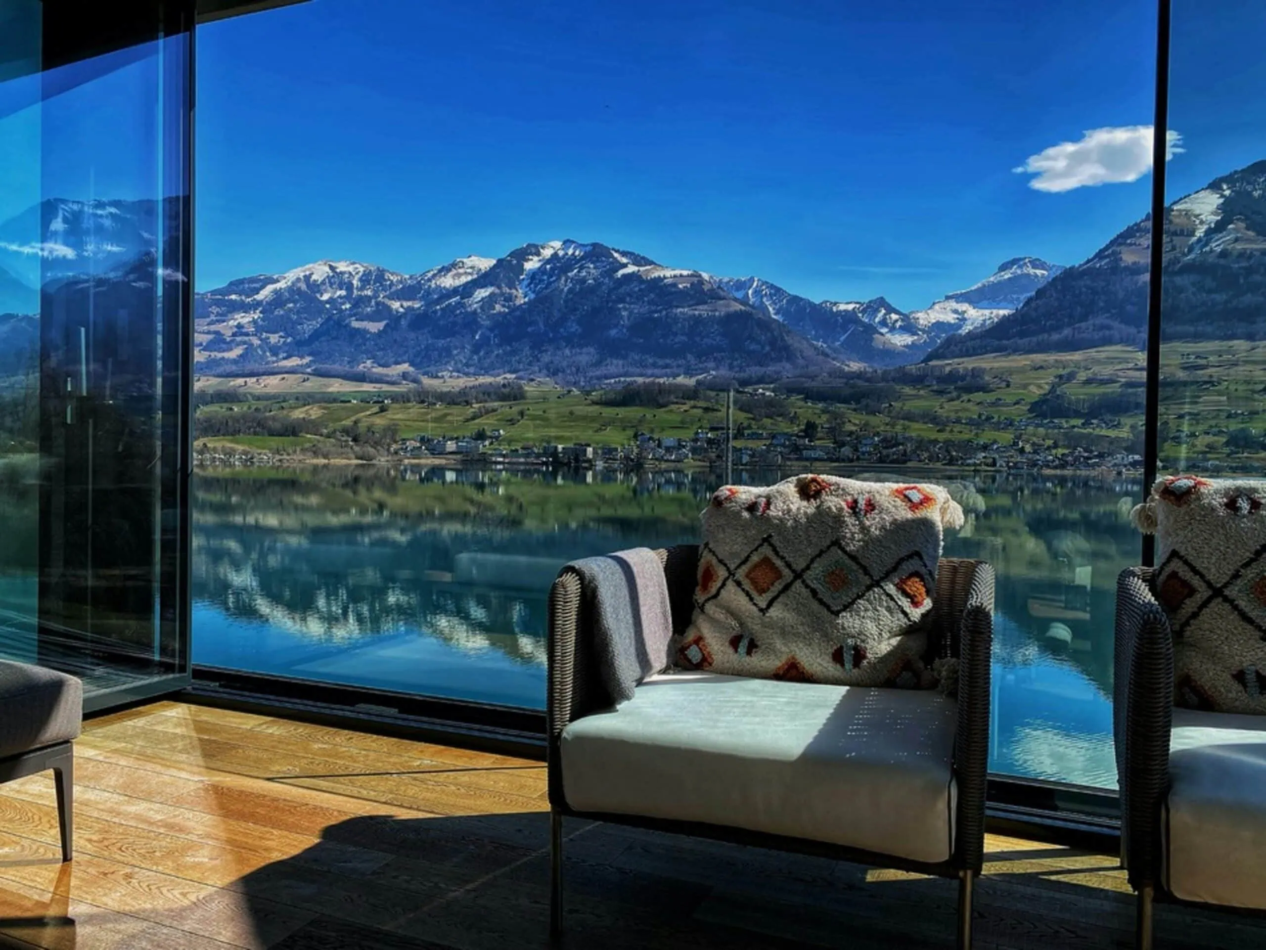 View from a modern interior through floor-to-ceiling windows onto a calm lake and snow-covered Alps